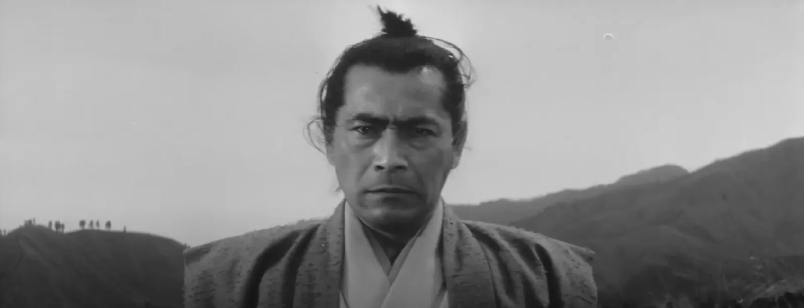 A lone samurai fixing an alert, poised stare, mountains and distant figures behind, in a scene from Masaki Kobayashi's Samurai Rebellion (1967)