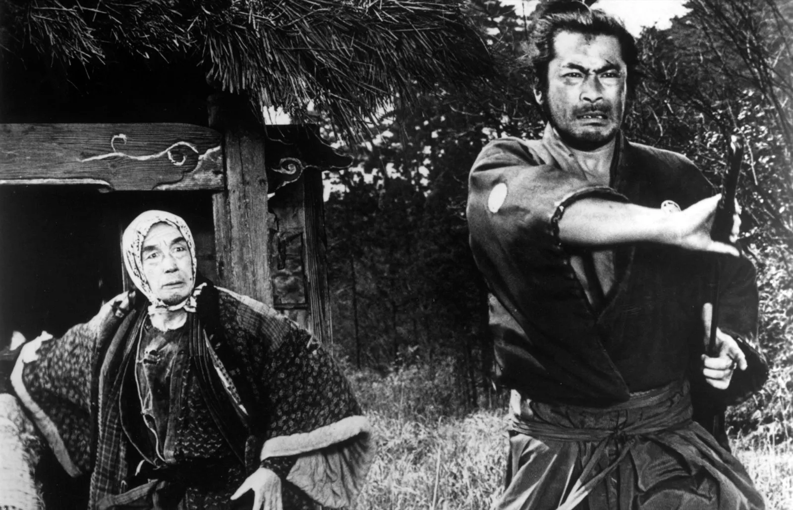 Toshiro Mifune as a samurai mid-strike, sword drawn across his body, an older woman seated beside him, in black and white