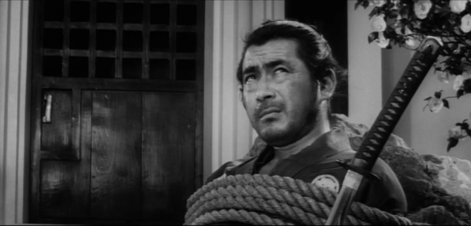 A weathered samurai seated with a coil of rope across his chest and a sword propped beside him, looking up to size up a new situation, camellias behind him