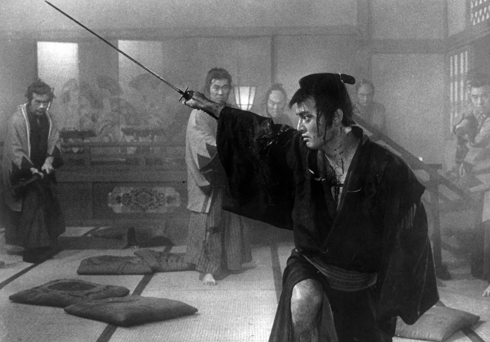 A blood-spattered swordsman kneels with his sword raised and pointed, surrounded by watching figures in a smoky room, in black and white