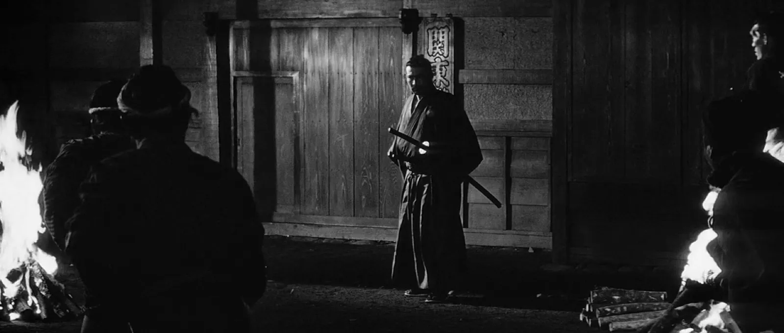 A lone samurai standing before a large closed wooden gate at night, flanked by fires, watched by figures in the dark, in black and white