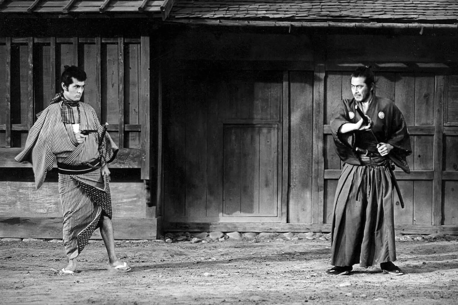 Two samurai facing off at a distance in a village street, one drawing a pistol, the other with his hand on his sword hilt, in black and white