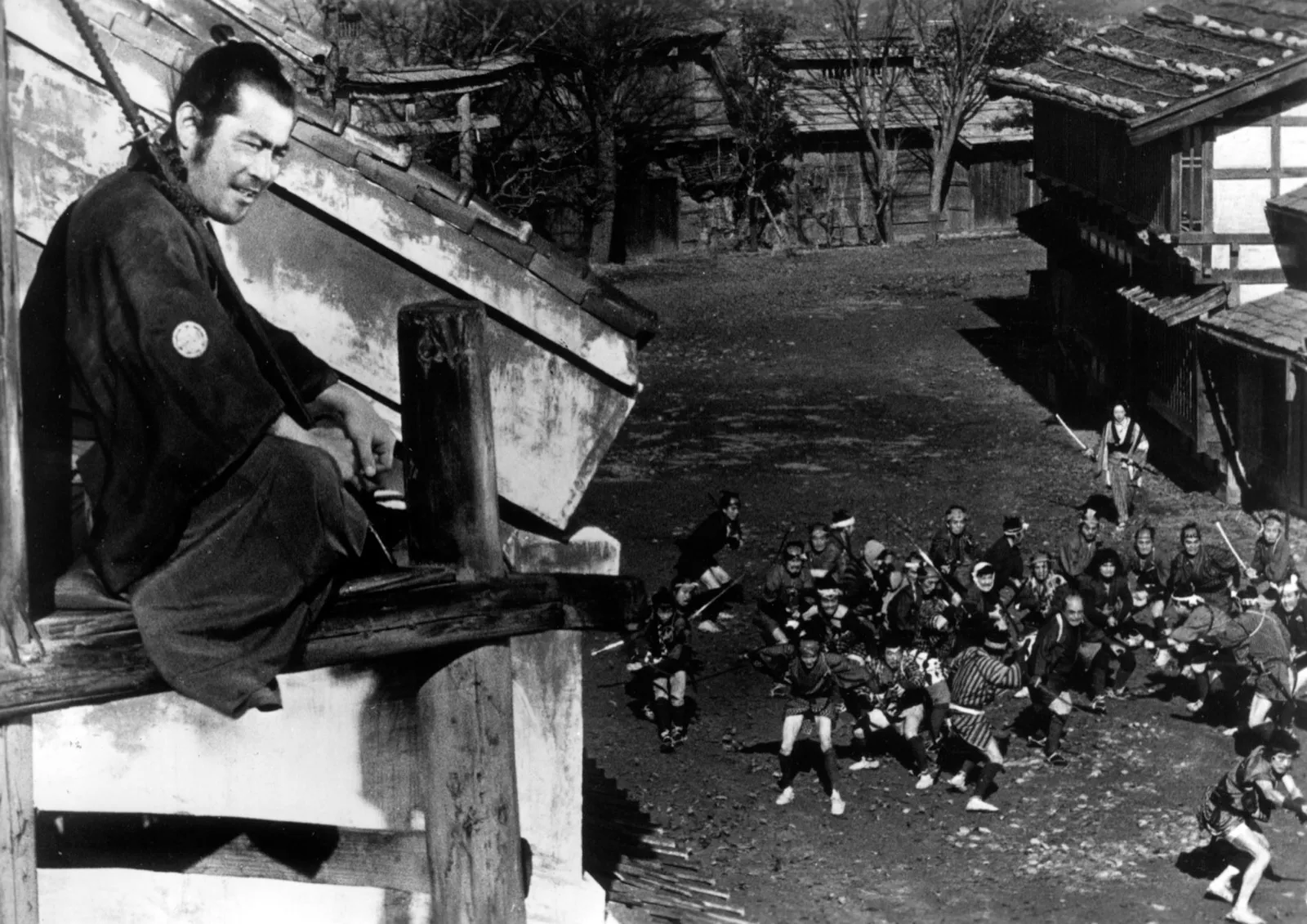 A lone samurai perched high on a wooden platform, watching a crowd of armed men gathered in the street below, in black and white